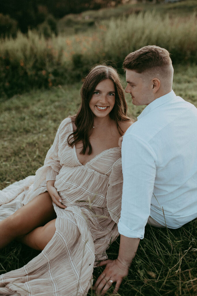 The expecting couple leans against each other, sitting on the grass at NCMA, Raleigh, NC.