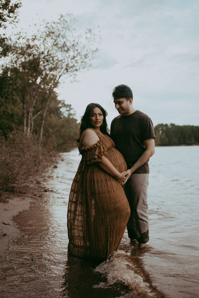 A glowing mom-to-be in a brown boho maxi dress stands on the rocky lakeshore, holding hands with her partner as the golden sunset reflects on the water.