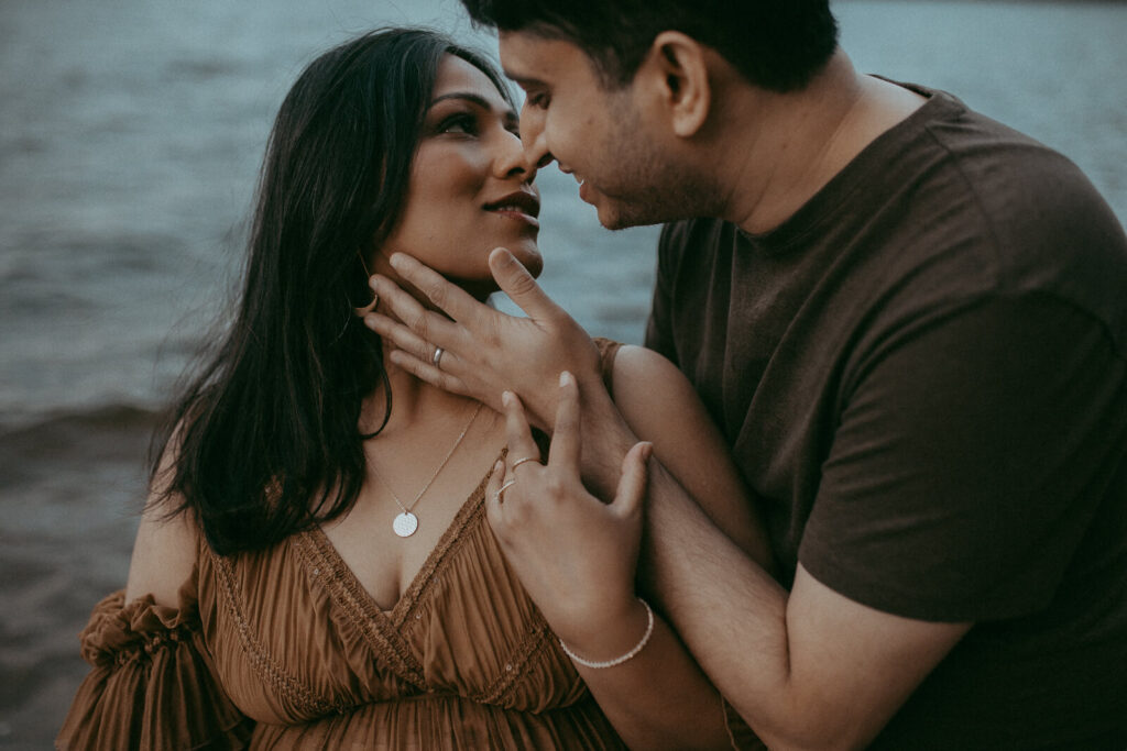 A serene maternity portrait of a mother-to-be in a brown boho dress, holding her bump while standing beside her partner on a peaceful lake shore.