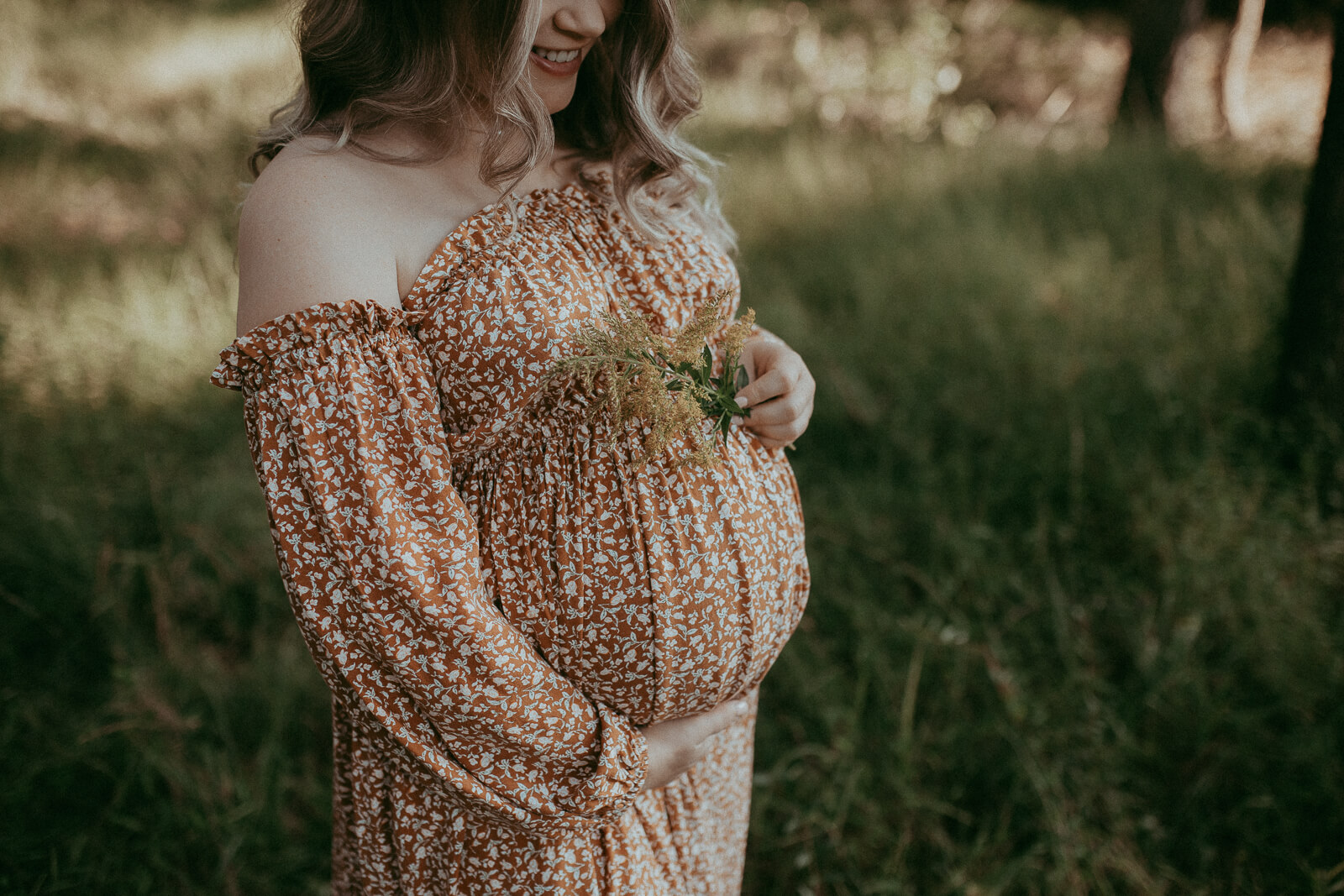 A close-up of mom-to-be’s hands wrapped around her belly and holds yellow flowers in her left hand, highlighting the delicate details of her floral dress and the joy of pregnancy.