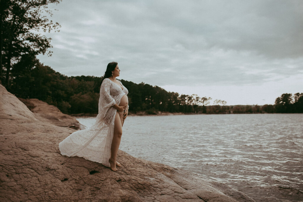 A timeless maternity portrait of a woman in a lace Reclamation gown, standing with confidence near Jordan Lake, her long black hair framing her face. The portrait was taken by Cary maternity photographer - Victoria Vasilyeva Photography.