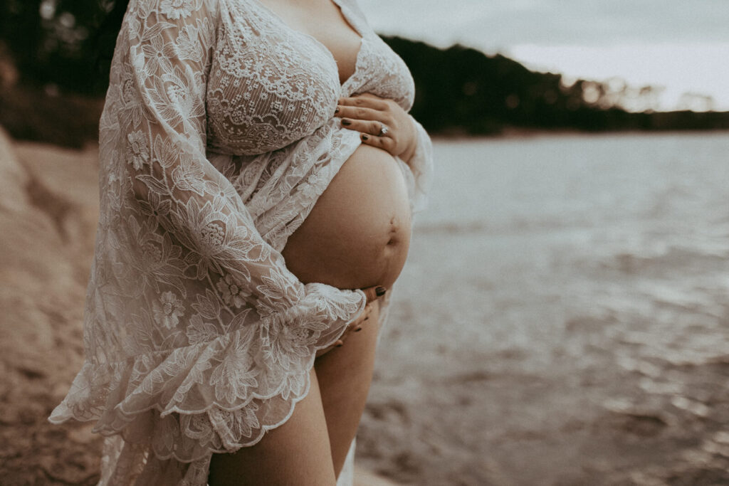 A radiant pregnant woman poses on a textured rock, wearing a dreamy lace maxi dress, gazing down at her belly with a peaceful expression.
