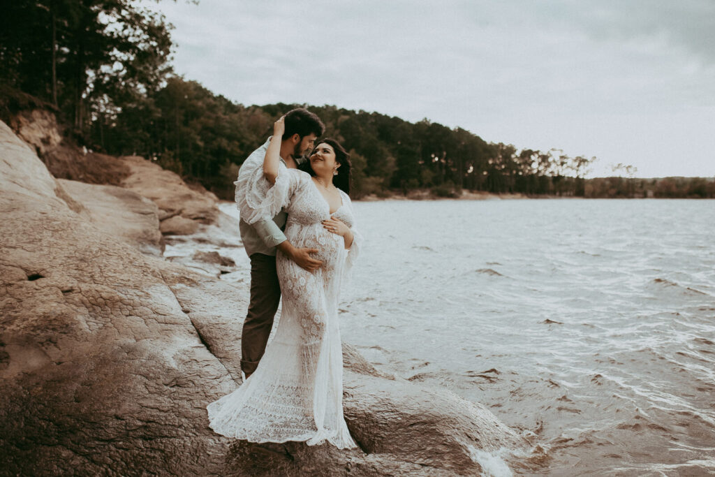 A stunning pregnant woman with long black hair in a flowing lace maxi dress stands by a serene lake, her partner holding her belly as they gaze at each other. The portrait was taken by Cary maternity photographer - Victoria Vasilyeva Photography.