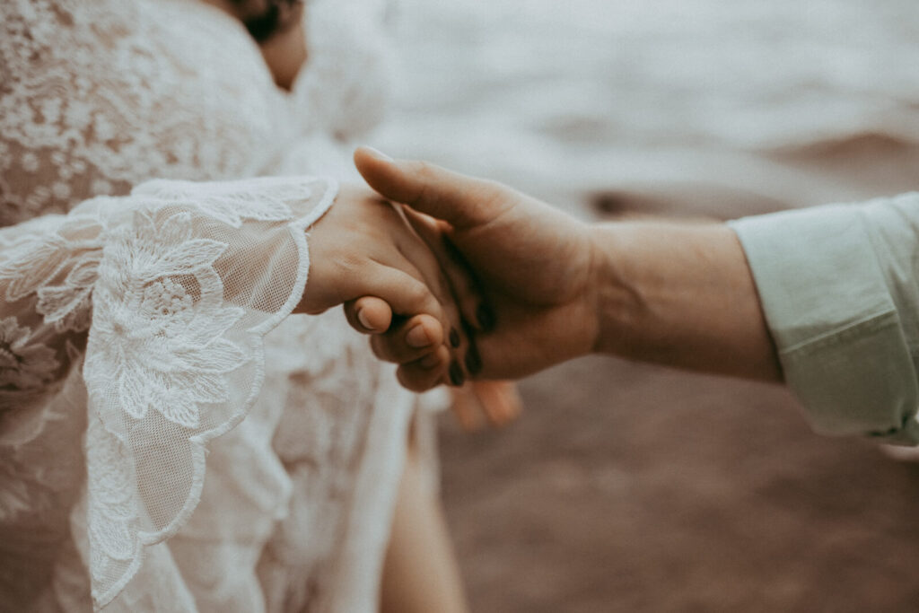 A glowing pregnant woman in a lace maxi dress leans into her partner’s touch, their hands clasped together as they stand on a rocky lakeshore, the soft wind adding movement to her gown.