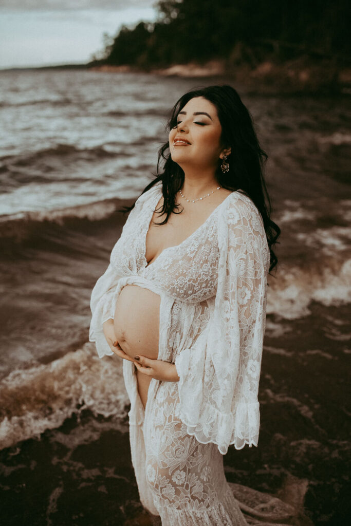 A stunning mother-to-be with long black hair cradles her baby bump while wearing a delicate lace maxi dress from Reclamation, bathed in golden light near the Cary Birth Center.