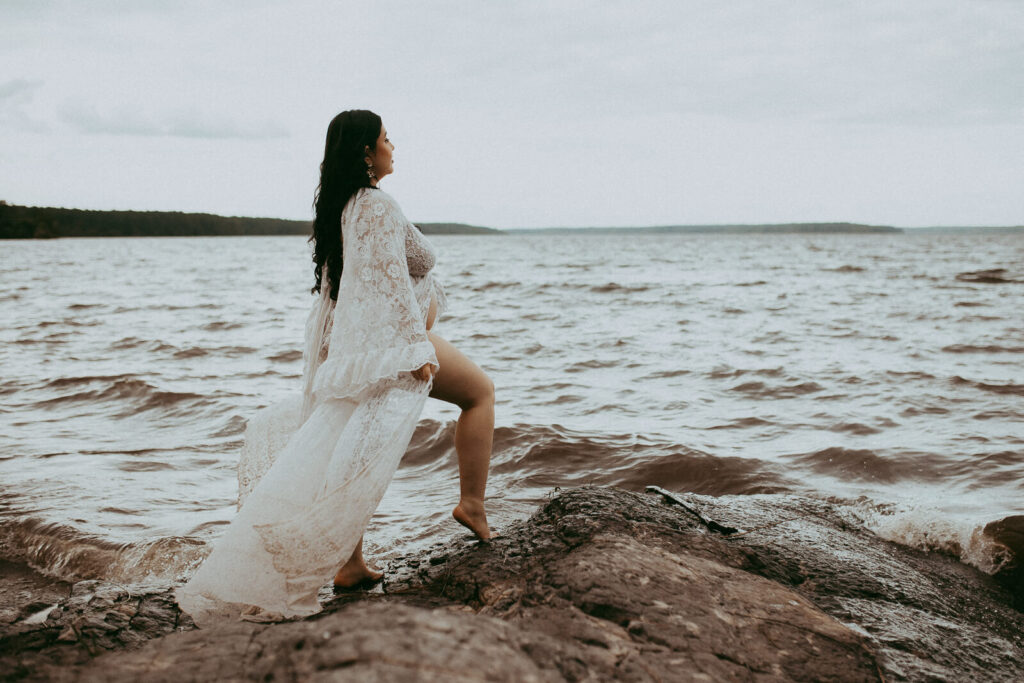 A mother-to-be in a delicate lace dress stands gracefully on a rock by the lake, her long black hair cascading down as she rests a hand on her sides. The portrait was taken by Cary maternity photographer - Victoria Vasilyeva Photography.