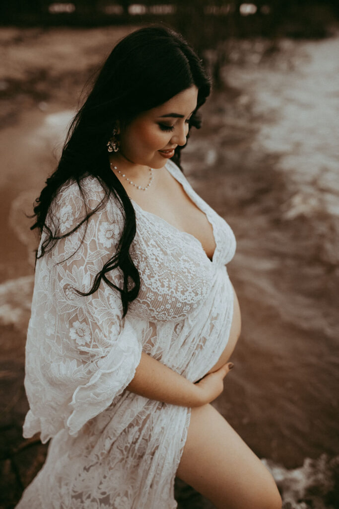 A glowing maternity client in an intricate lace Reclamation dress gently holds her belly, looking down with a soft smile as the sun sets near Cary Birth Center.