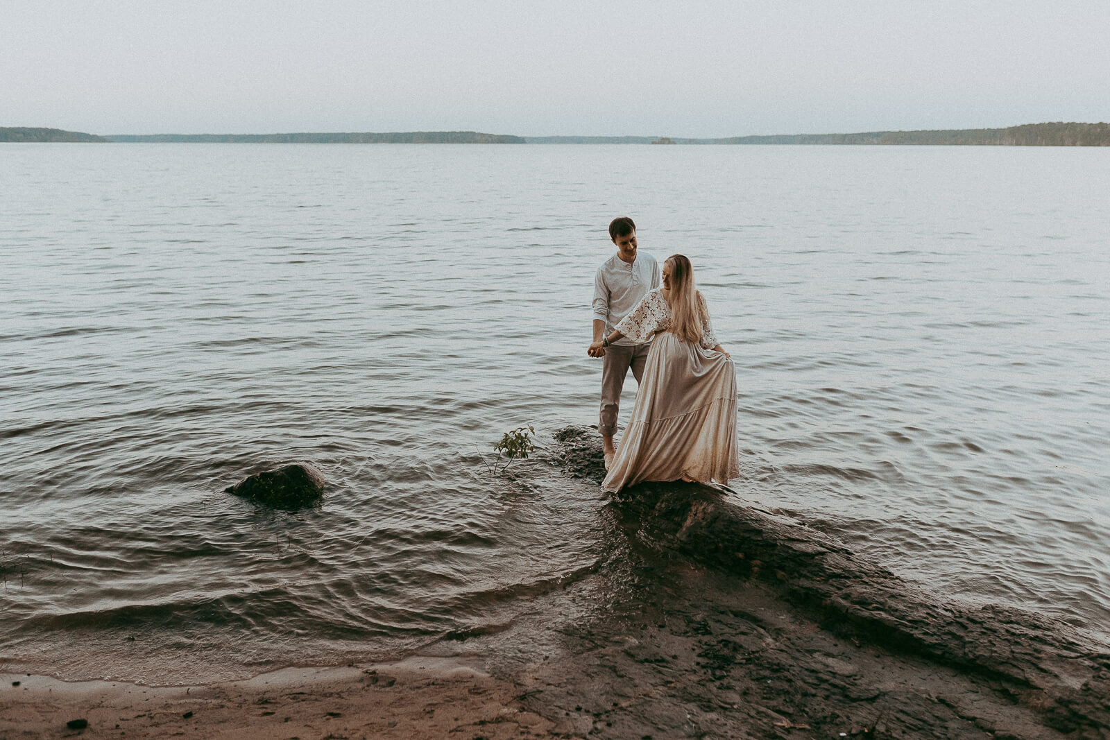 Expectant parents walking hand-in-hand along a path at sunset. Mom with long blonde hair wears a boho crochet top and linen maxi skirt, Dad is in light casual attire. Photo by Victoria Vasilyeva Photography.