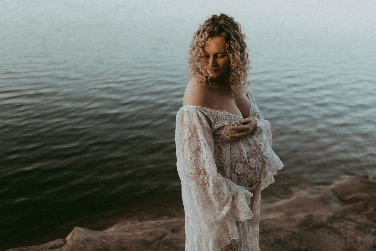 Stunning mom-to-be is staying on a rock near the lake shore. She looks down at her shoulder and hugs the belly. The portrait was taken near Bull City Beginnings by Victoria Vasilyeva Photography - Durham maternity photographer.