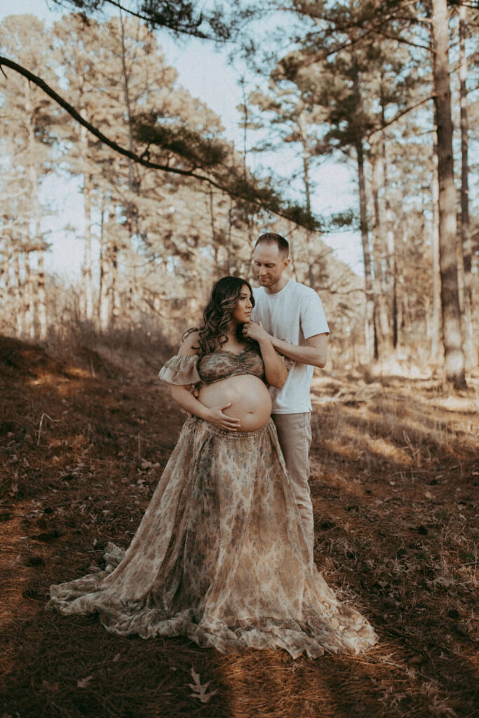 A pregnant woman in a flowing, floral-patterned dress from Reclamation Brand stands in a pine forest with her partner. She gently cradles her bare belly while he stands behind her, holding her close and tenderly touching her face. The warm, earthy tones of the forest and soft natural light create a serene and intimate atmosphere. Maternity photo session took place in Raleigh by Victoria Vasilyeva Photography.