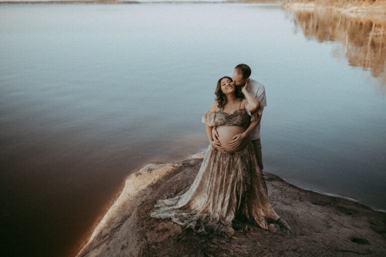 In soft, golden hour light, a couple shares a tender embrace by a still lake. The expectant mother wears a sheer, boho-style dress, her partner standing behind her with his arms wrapped around her, conveying warmth and affection. The portrait was taken by Victoria Vasilyeva Photography during maternity photo session in Raleigh, NC.