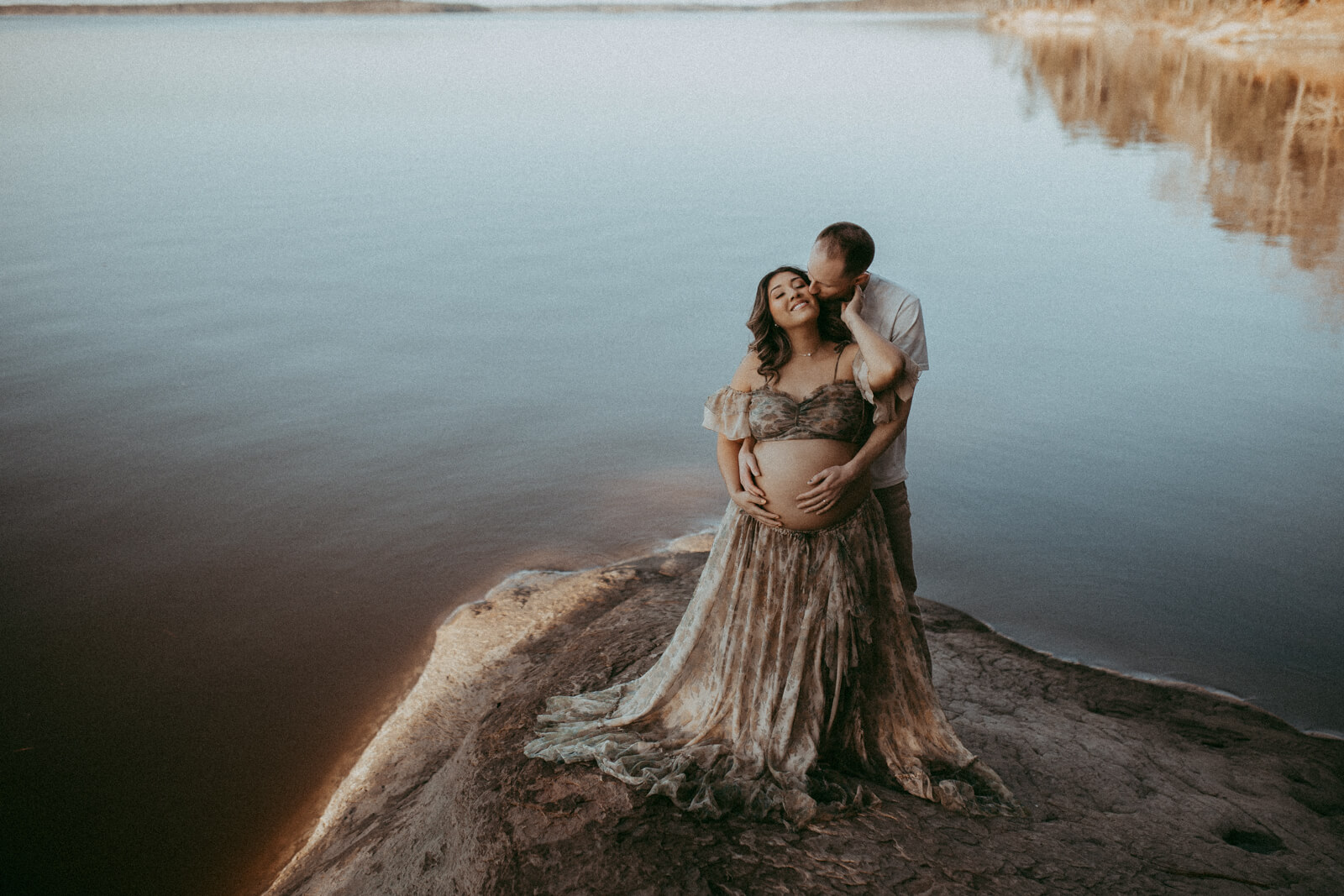 In soft, golden hour light, a couple shares a tender embrace by a still lake. The expectant mother wears a sheer, boho-style dress, her partner standing behind her with his arms wrapped around her, conveying warmth and affection. The portrait was taken by Victoria Vasilyeva Photography during maternity photo session in Raleigh, NC.