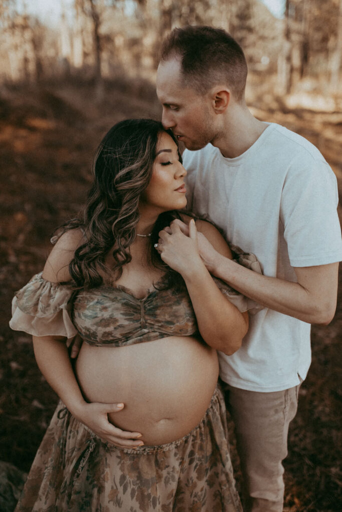 A pregnant woman in floral-patterned set from Reclamation Brand stands among high trees with her partner. She gently cradles her bare belly while he stands om her left side, holding her close and tenderly touching her face. Maternity photo session took place in Raleigh near Davis Family Chiropractic office by Victoria Vasilyeva Photography.