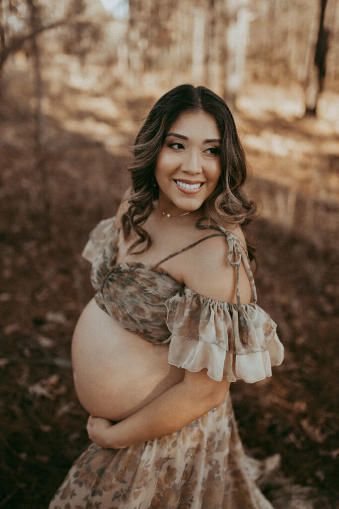 A pregnant woman in floral-patterned set stands among high trees. She gently hugs her belly while looks at left side. Maternity photo session took place in Raleigh near Davis Family Chiropractic office by Victoria Vasilyeva Photography.