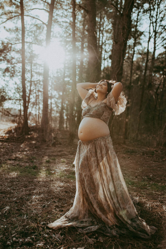 A pregnant woman stands in a sun-dappled forest, bathed in warm golden light filtering through the trees. She wears a flowing, earthy-toned, off-the-shoulder top and skirt that accentuate her bare baby bump. With her hands resting behind her head and a soft smile on her face, she appears peaceful and radiant, embodying serenity and strength in a natural woodland setting. The portrait was taken by Victoria Vasilyeva Photography during maternity photo session in Raleigh, NC.