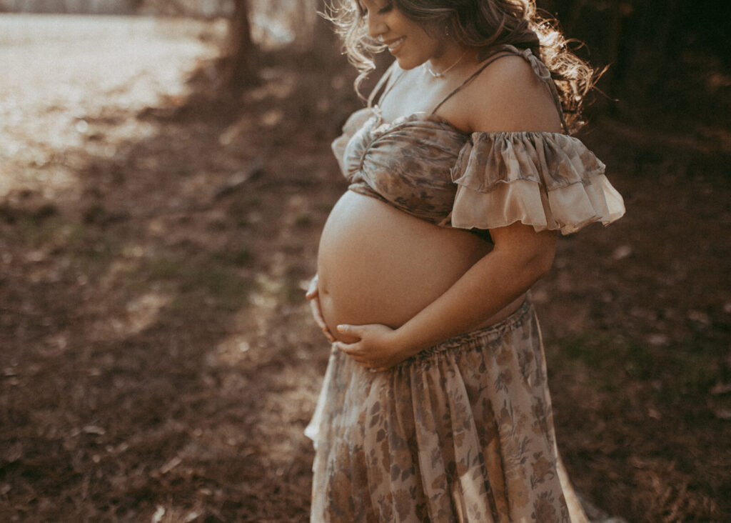 A pregnant woman stands in a sun-dappled forest, bathed in warm golden light filtering through the trees. She wears a flowing, earthy-toned, off-the-shoulder top and skirt that accentuate her bare baby bump. With her hands resting on her belly, she appears peaceful and radiant, embodying serenity and strength in a natural woodland setting. The portrait was taken by Victoria Vasilyeva Photography during maternity photo session in Raleigh, NC.
