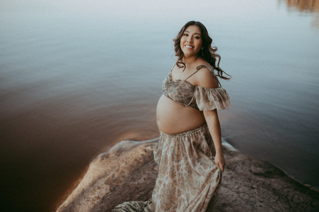 A pregnant woman stands on a rocky lakeshore in a flowing, floral gown, smiling at the camera. The calm water and muted tones create a peaceful, romantic setting. The portrait was taken by Victoria Vasilyeva Photography during maternity photo session in Raleigh, NC.