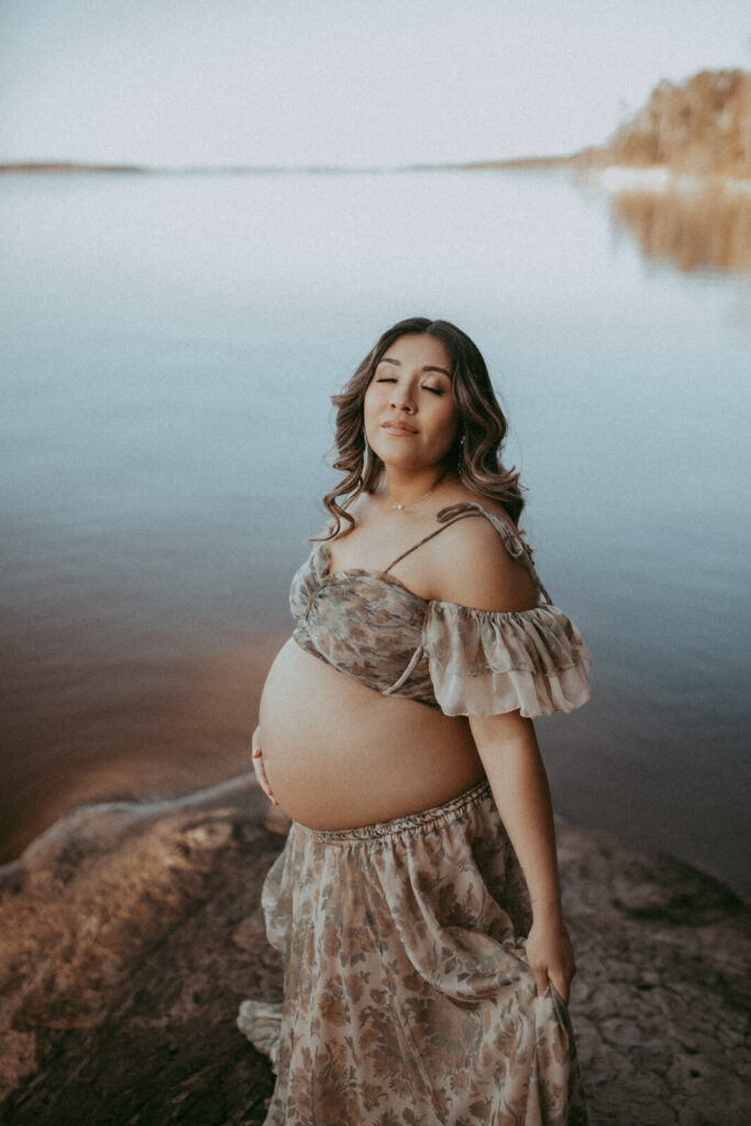 A pregnant woman stands on a rocky lakeshore in a flowing, floral gown, facing to the camera. The calm water and muted tones create a peaceful, romantic setting. The portrait was taken by Victoria Vasilyeva Photography during maternity photo session near Davis Family Chiropractic in Raleigh, NC.