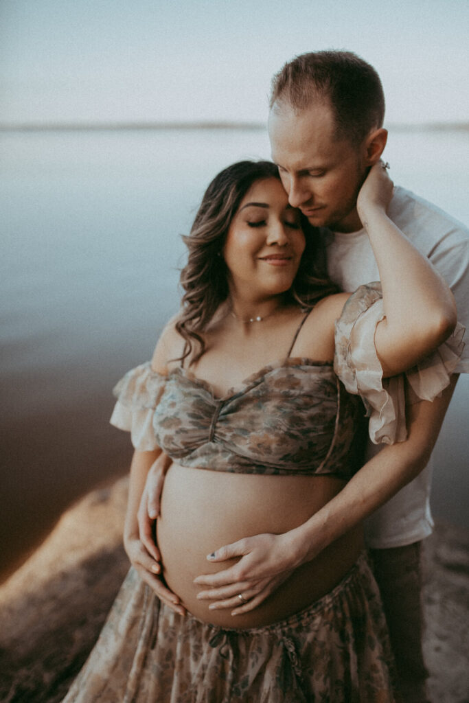 A maternity couple poses on the edge of a tranquil lake, with the woman’s bare belly cradled in her partner’s hands. She wears a long, ethereal dress while he gently kisses her, capturing an intimate and serene moment. The portrait was taken by Victoria Vasilyeva Photography during maternity photo session near Davis Family Chiropractic in Raleigh, NC.