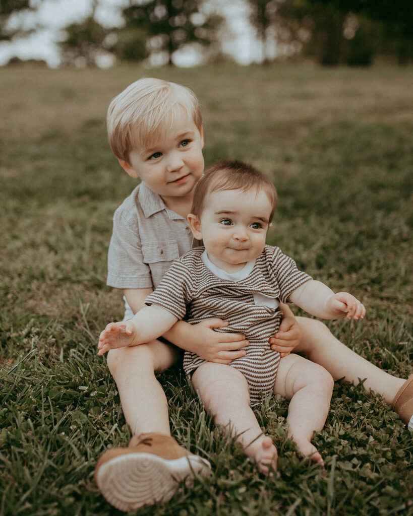 2 brothers with blonde hair, wearing stripped outfits, sit on a grassy hill at NCMA in Raleigh, NC. The boys are happy and smiling at the parents.