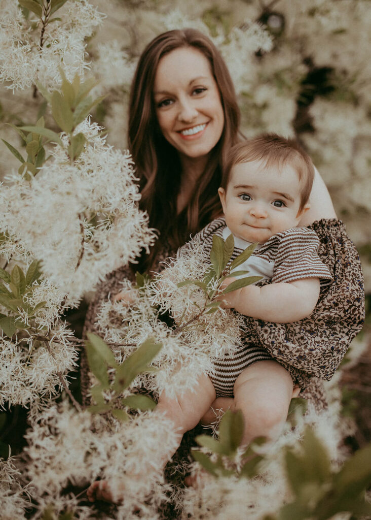 A smiling woman with long brown hair, wearing a dark patterned dress, stays between blooming trees with a baby boy in her arms. The boy, with blonde hair and a striped romper, is also smiling at the camera. Family photo session in Raleigh, NC by Victoria Vasilyeva Photography.