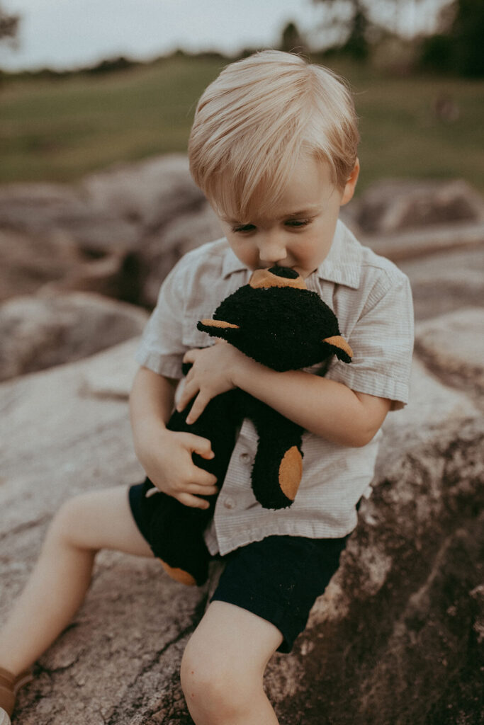 3-years old blonde boy sits on large rocks at NCMA in Raleigh, looking down at black teddy bear and kissing him. Family photo session in Raleigh, NC by Victoria Vasilyeva Photography.