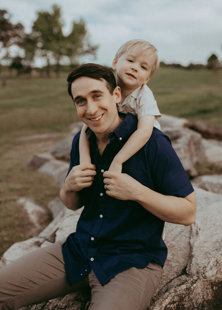 A father holds his son on his back. Both are smiling to the camera with Victoria Vasilyeva Photography - Raleigh family photographer.