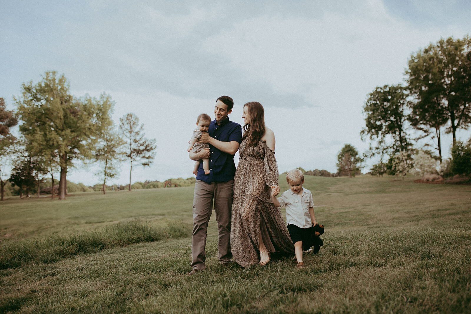 A family of four walks across a green field with trees in the background at NCMA. A man carries a baby boy, a woman in a maxi dress walks beside him, and a young boy holds her hand and black teddy bear. Family photo session was done after the family attended infant swim lessons in Raleigh, NC