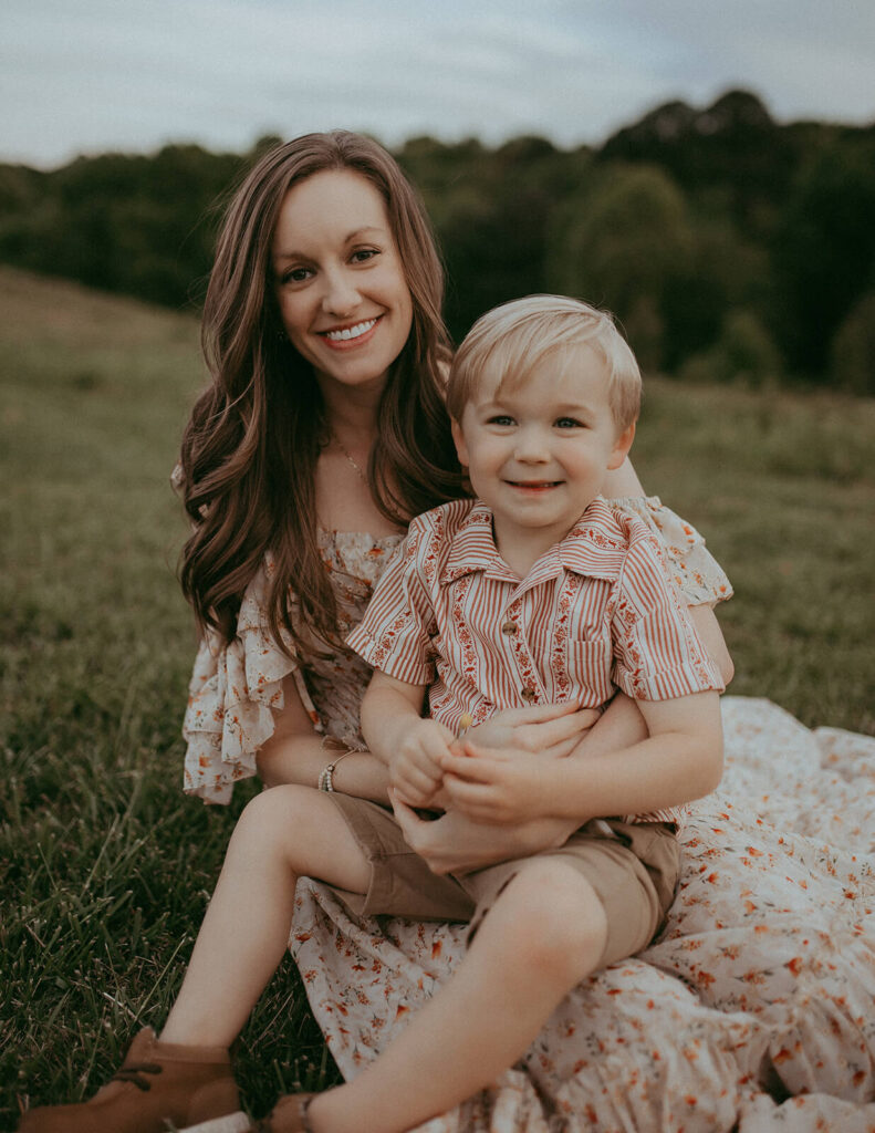 A smiling woman with long brown hair, wearing a floral dress with raffles, sits on a grassy hill at NCMA with her 3 years old son sitting in front of her. The boy, with blonde hair and a striped shirt and tan shorts, is also smiling at the camera. Family photo session was done after the family attended infant swim lessons in Raleigh, NC