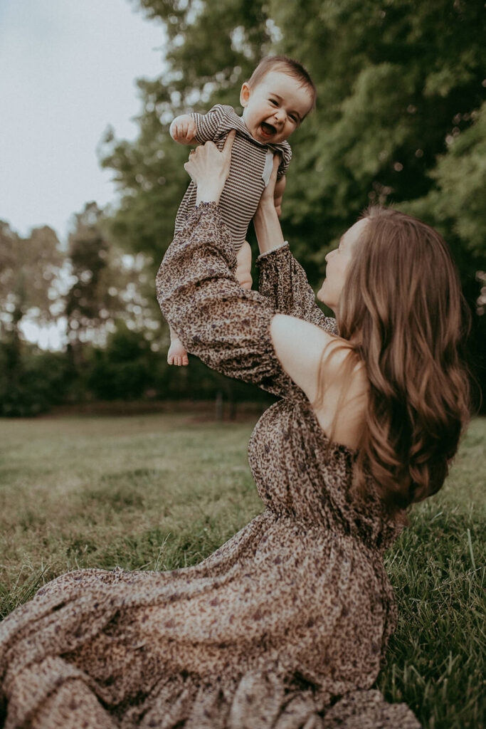 Side view of a gorgeous woman with long wavy hair. She is sitting on the ground and lifting a baby dressed in a striped romper. Both appear joyful amidst trees and grass. Family photo session took place in Raleigh, NC by Victoria Vasilyeva Photography.