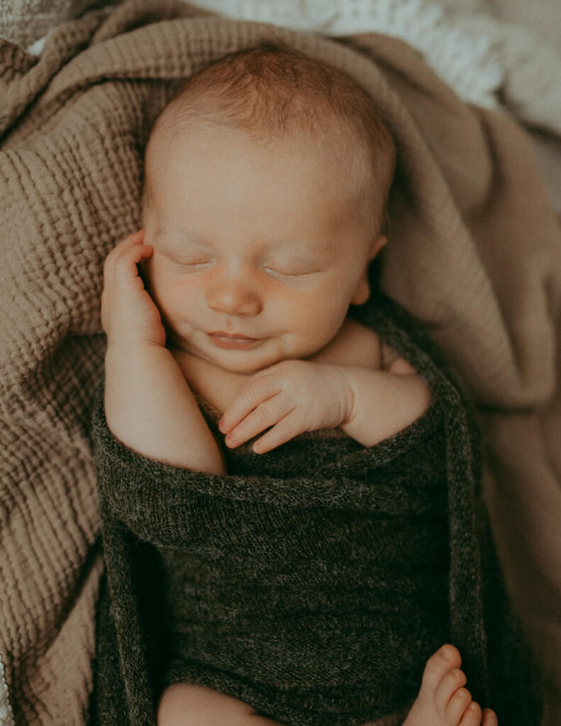 Close-up, overhead view of a newborn baby boy sleeping soundly, swaddled in a soft dark blanket. This image evokes the peaceful outcomes often sought by parents utilizing Lactation Consultant services in Fayetteville, NC