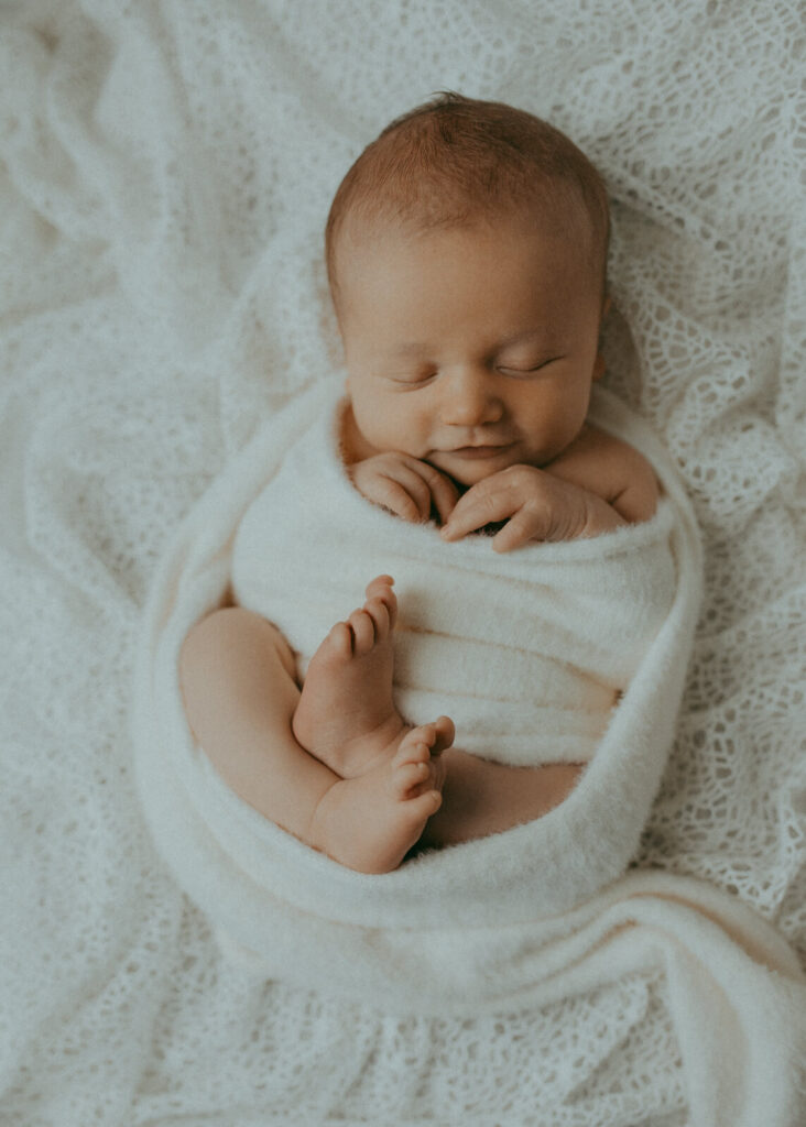 A tranquil portrait of a swaddled infant, highlighting their delicate features and tiny hands and feet, against a soft white background. This portrait was taken by newborn photographer in Fayetteville, NC - Victoria Vasilyeva Photography.