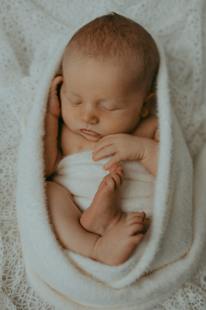 10 lbs newborn baby boy, swaddled in a white, soft blanket, sleeps soundly with their eyes closed and tiny hands resting near their face, one foot peeking out. Newborn photo session in Fayetteville, NC with Victoria Vasilyeva Photography.