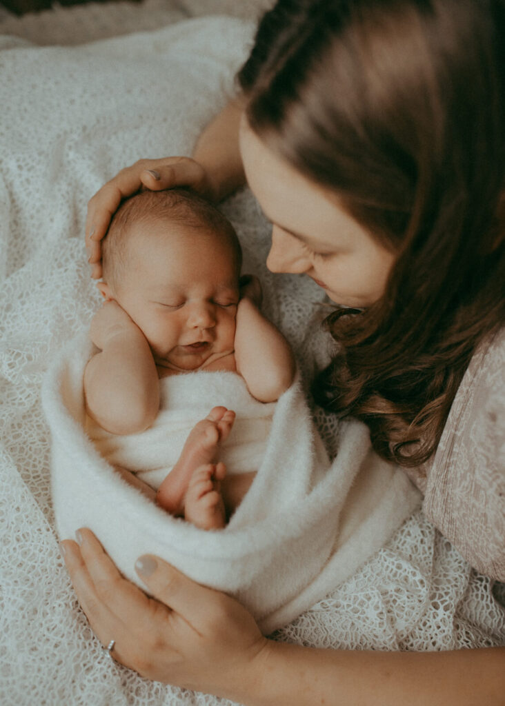 10 lbs newborn baby boy wrapped in white cozy blanket. His mom gentle hugs him and smiles down. Newborn photo session in Fayetteville, NC with Victoria Vasilyeva Photography.
