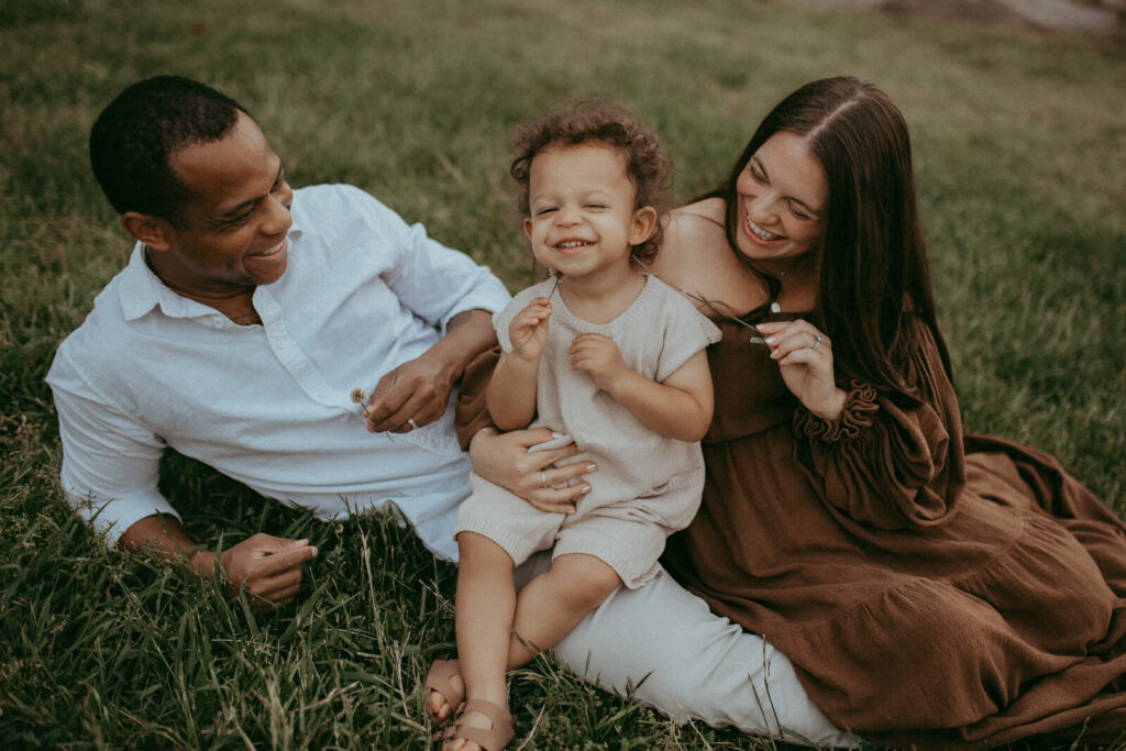 A heartwarming moment between mom, dad and the child outdoors. The child sits on the dad's legs and smiling at the camera, all looking happy. Family photo session by Raleigh family and newborn photographer - Victoria Vasilyeva Photography.