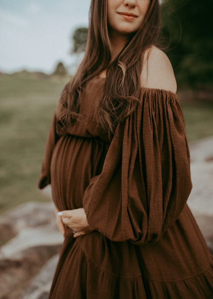 The portrait of pregnant woman in boho brown dress. She gently hugs her belly and turns her face to her left shoulder.