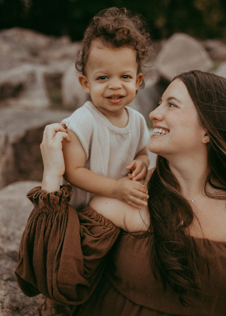 A joyful mother looks up at her smiling toddler, who is perched on her shoulders, against a blurred background of large rocks. He wears cute knitted overoll from Lora Belle Baby Store. Family portrait session was taken by Raleigh family and newborn photographer.