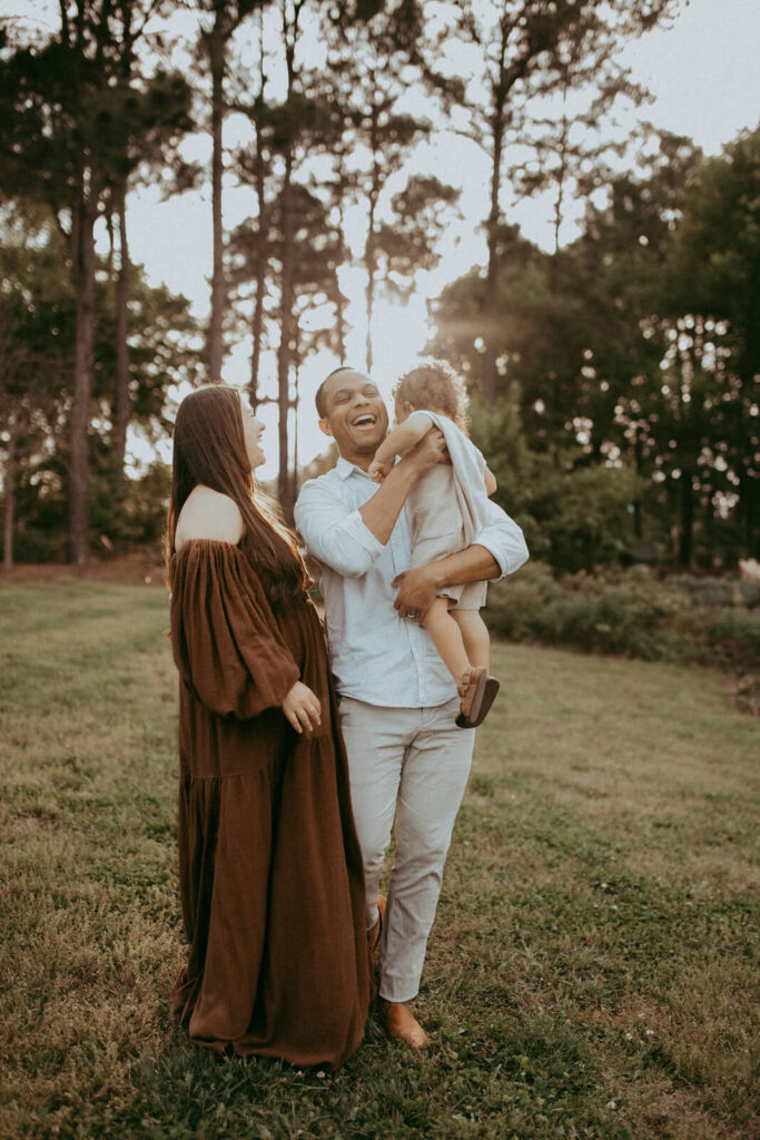 A family of three stands in a grassy field. A dad with a light-colored shirt and pants holds a baby boy with curly hair, while a woman in a long brown dress stands nearby, looking towards the child. A toddler wear knitted romber from Lora Belle Baby boutique. Family photo session by Victoria Vasilyeva Photography.