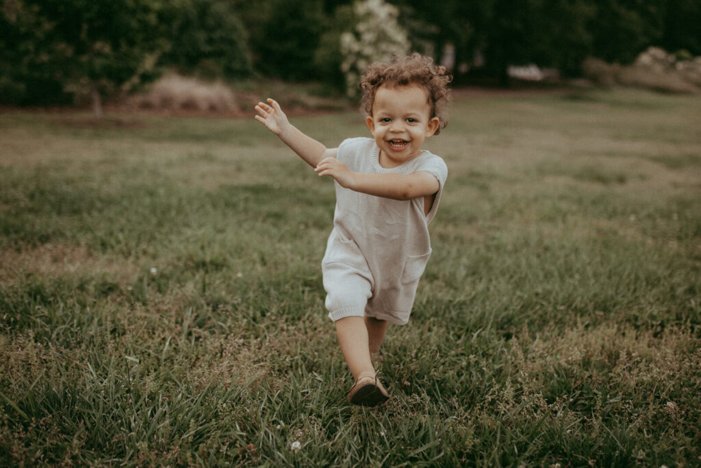A portrait of a smiling toddler running toward camera. He has curly hair and wear tan knitted overall from Lora Belle Baby store. The portrait was taken by Raleigh family and newborn photographer - Victoria Vasilyeva Photography.
