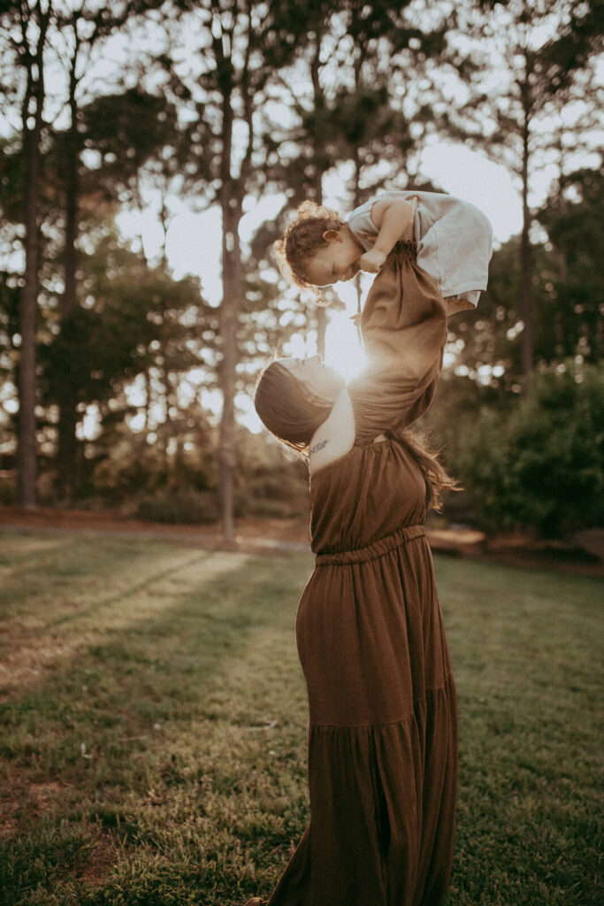 The portrait of a happy mother and her young child. The child, with curly hair, laugh at her while the mother, in a brown maxi dress, smiles up at the child. Family portrait was taken by Raleigh family and newborn photographer.