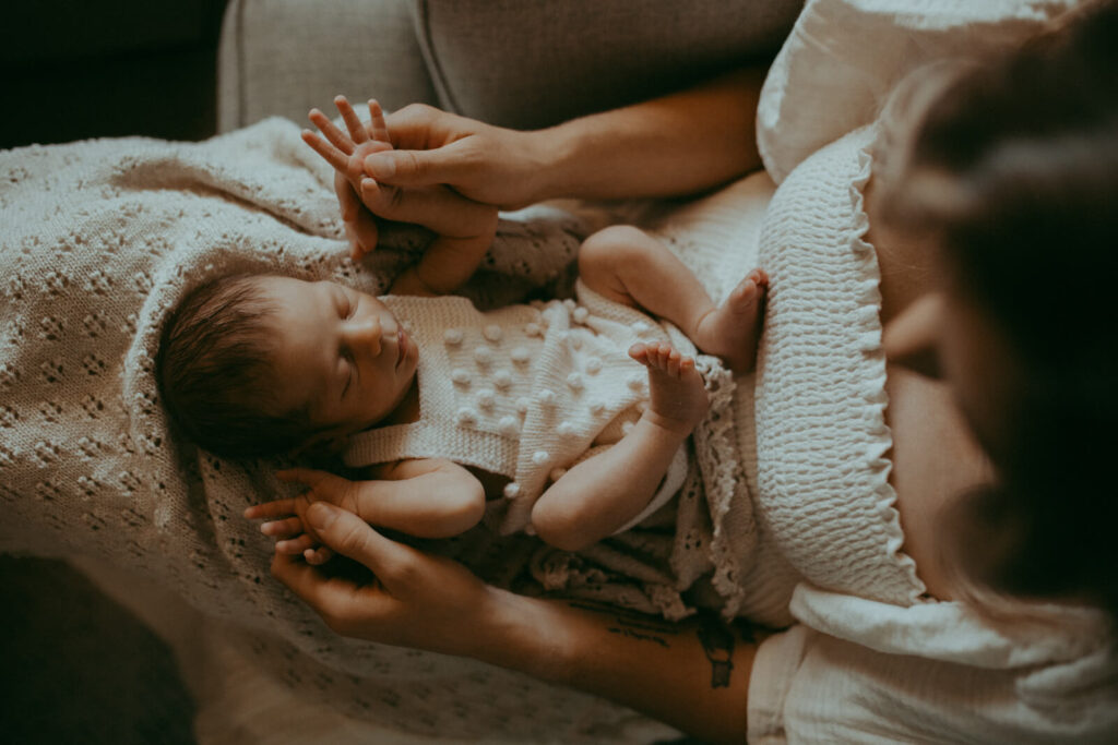 An overhead shot captures a mom cradling a sleeping newborn baby boy in her arms. The newborn, wearing a white knitted romper with textured dots, is snuggled on a light-colored, knitted blanket. A mom is wearing a off-white ruffled dress. The lighting from the window is soft and warm, creating a tender and intimate atmosphere. Newborn portrait session with Raleigh newborn photographer. Night nurse in Raleigh, NC