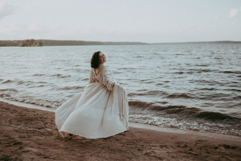 Expecting woman embracing by a lake. She wearing a white linen maternity dress and smiling back at the camera.