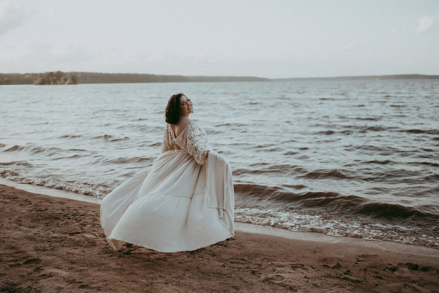 Expecting woman embracing by a lake. She wearing a white linen maternity dress and smiling back at the camera.