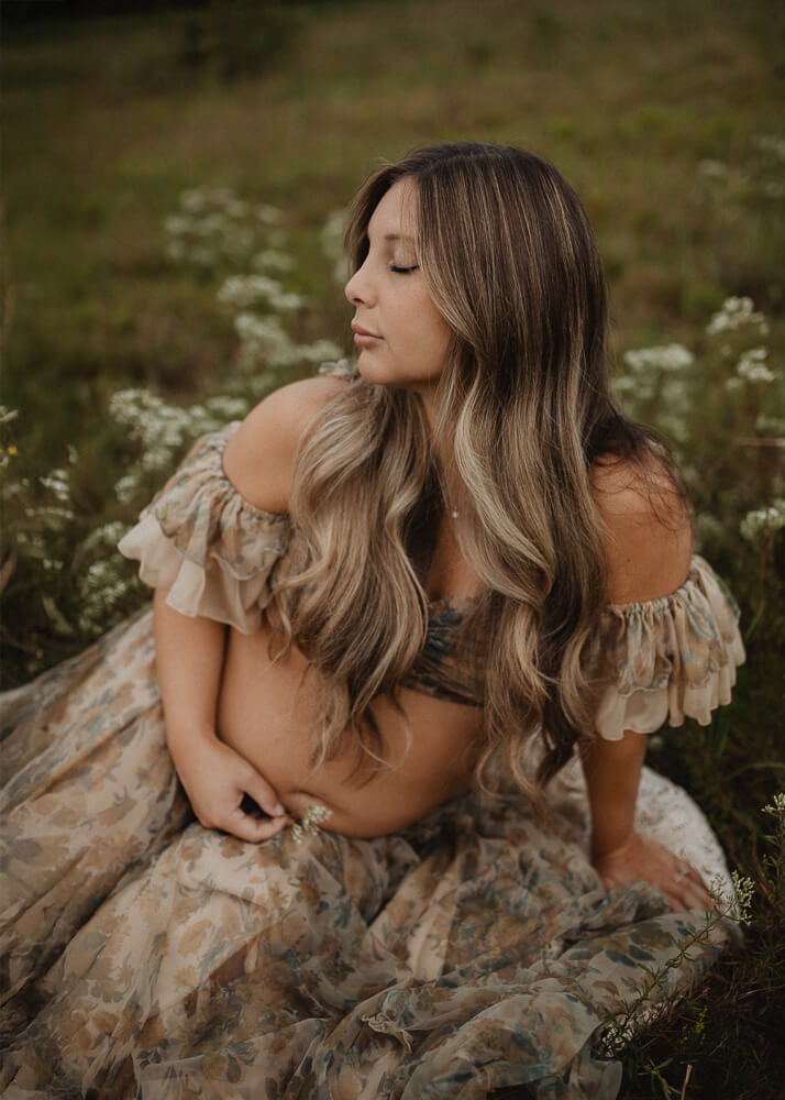 Pregnant woman sitting in a field of wildflowers, holding her belly and wearing a floral gown during a maternity session. Read my blog about 4D Ultrasound Raleigh NC.