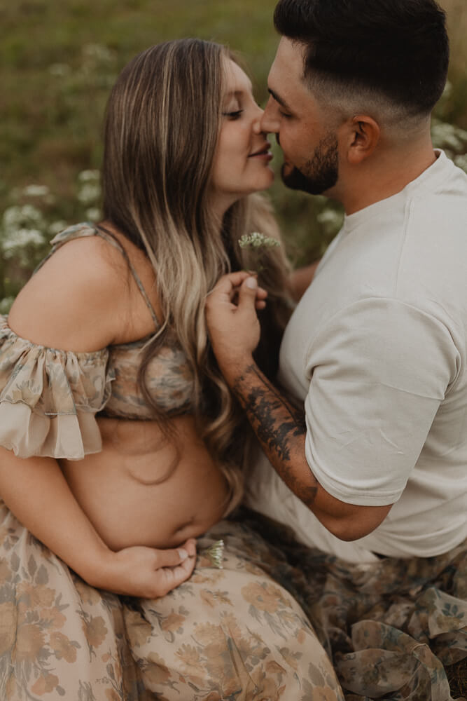 Expecting couple sharing a soft kiss in a grassy field, the mother-to-be holding her belly while wearing a flowing floral dress. Image featured in a 4D Ultrasound Raleigh NC article.
