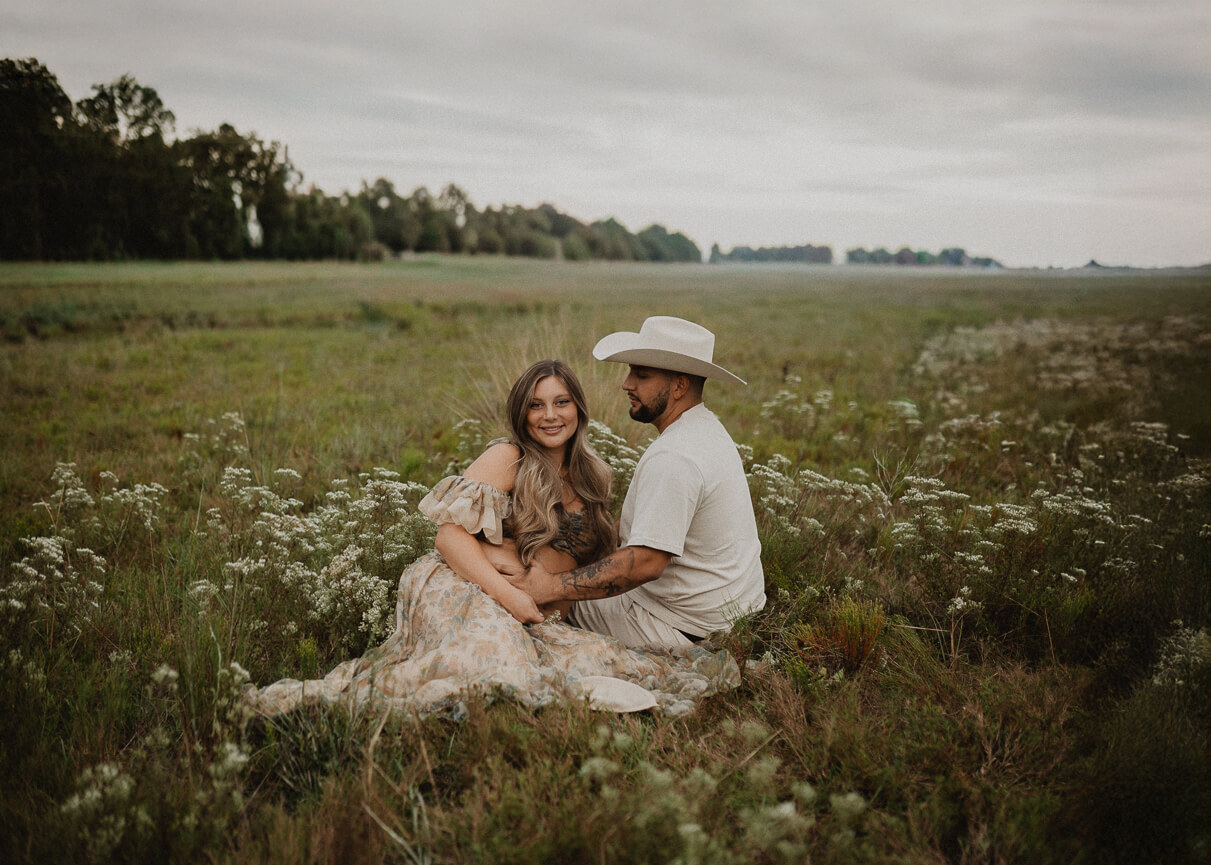 Expecting couple sitting together in an open field surrounded by wildflowers, smiling during an outdoor session with maternity photographer. Read my blog about 4D Ultrasound Raleigh NC.