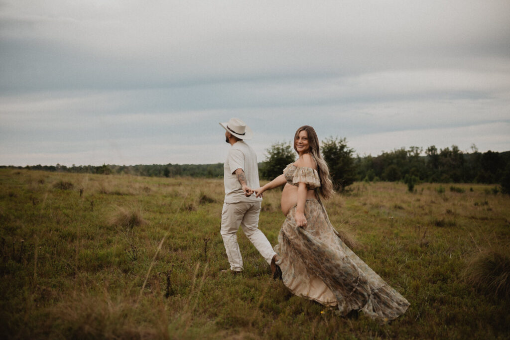 Pregnant woman with long hair holding her partner’s hand while walking through a wide open field in a floral maternity gown. Maternity photo session by Victoria Vasilyeva Photography.