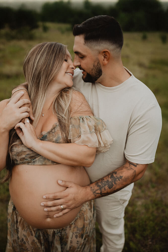 Close-up of an expecting couple embracing in a grassy field, highlighting the mother’s baby bump and flowing dress. Maternity photo session in Raleigh by Victoria Vasilyeva Photography.