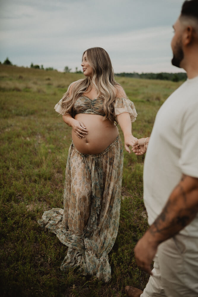 Pregnant woman holding her belly while walking through a natural field, wearing a long floral skirt and top from Reclamation brand. Maternity photo session in Raleigh by Victoria Vasilyeva Photography.