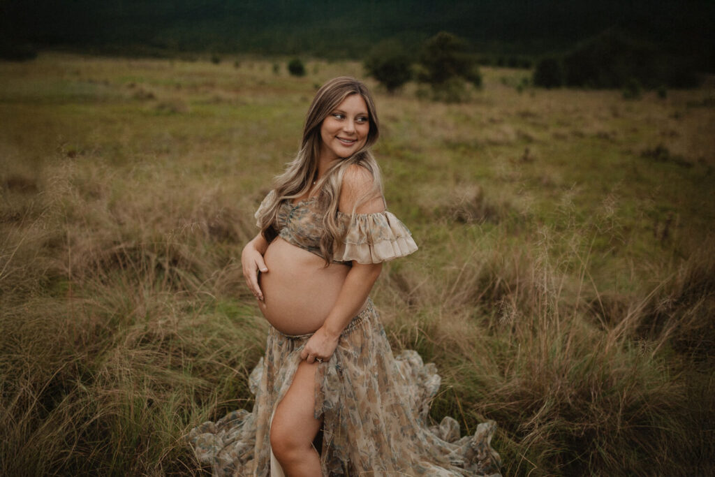 Expecting mother standing in tall grass, smiling and holding her belly during a golden-hour maternity session. Photo session took place in Raleigh, NC.
